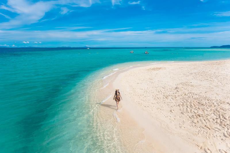 A tourist walking along a sandy beach with clear blue waters in Thailand, surrounded by a peaceful n